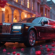 A glossy black Rolls-Royce stretch limousine is parked on a softly lit city street at twilight, surrounded by scattered red rose petals on the wet pavement. Ornate arches of a grand building glow warmly in the background, creating a romantic and luxurious ambiance.