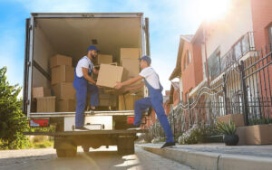 Two men in blue overalls are loading boxes into a moving truck in a sunny outdoor setting.