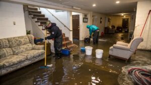 Two workers wearing waterproof gear clean up a heavily flooded basement, using brooms, buckets, and hoses to remove standing water around soaked furniture, including sofas and chairs.