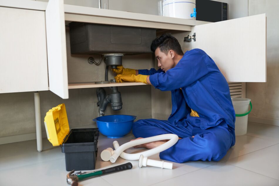 A professional plumber in blue overalls unclogging a kitchen sink drain under the cabinet using tools, representing expert drain cleaning services in Montreal.