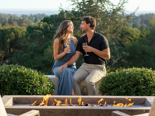 Couple in formal attire toasting wine at an outdoor firepit with forest and bay views at Chaminade Resort