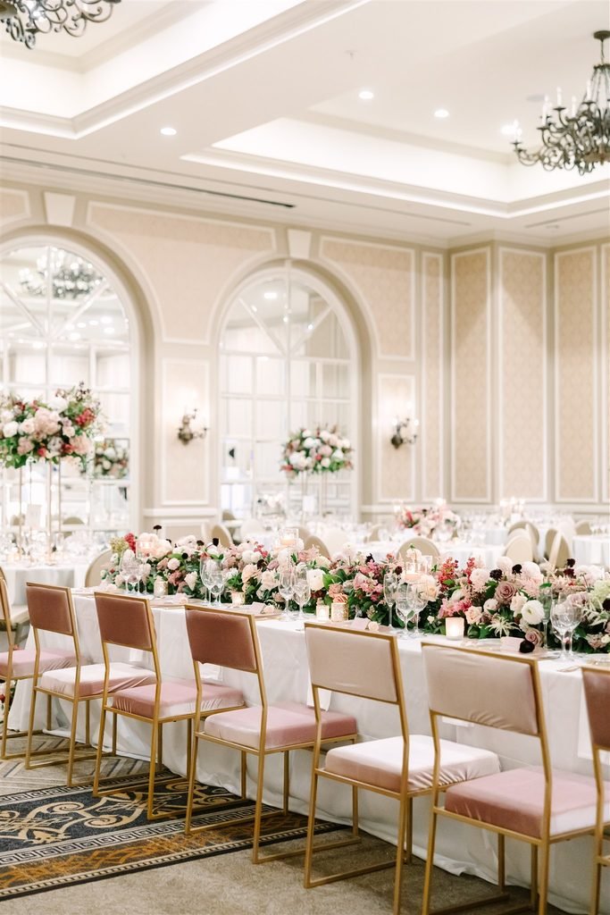 Long reception table with pink velvet chairs and a low floral runner beneath chandeliers and arched windows in a Dallas hotel ballroom at The Adolphus.