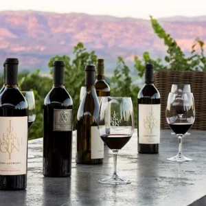 John Anthony wine bottles and glasses of red wine on an outdoor table with vineyard hills in the background